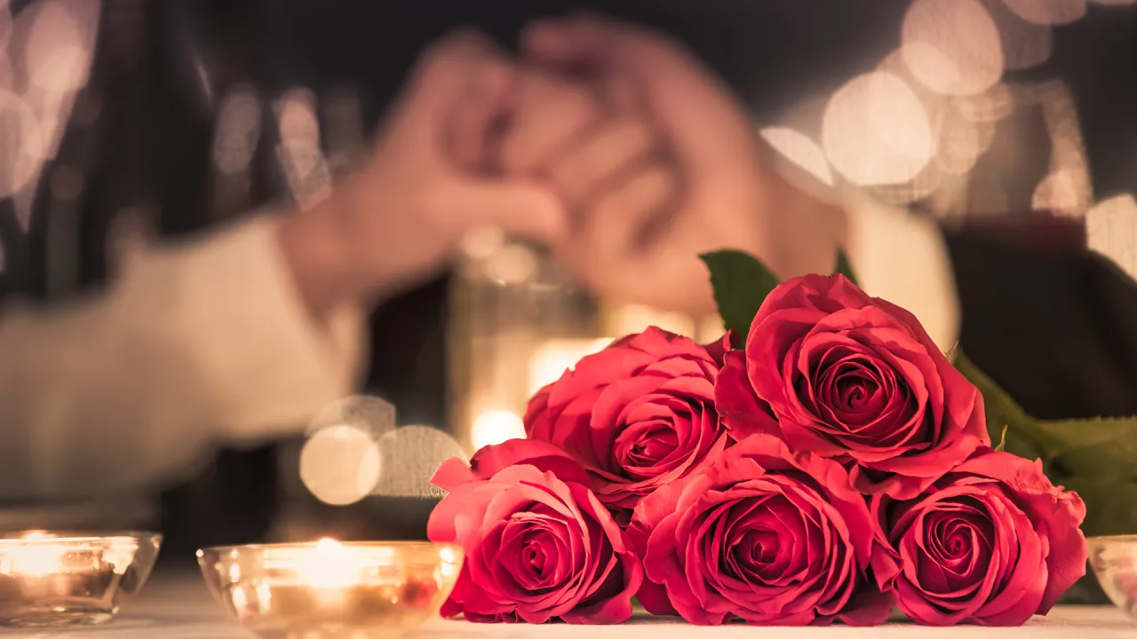 Bouquet of five red roses on a table with lit candles and two people holding hands blurred in the background.
